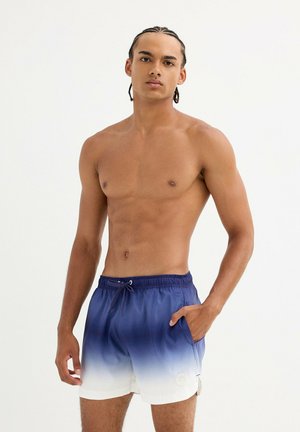 Joven con cabello trenzado que lleva pantalones cortos de baño en degradado azul y blanco, de pie con una mano en el bolsillo frente a un fondo liso.