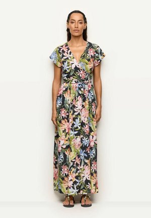 Femme debout portant une robe portefeuille noire à fleurs, manches courtes, et des sandales sur un fond uni blanc cassé.