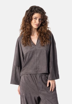 Femme aux longs cheveux bouclés portant un haut gris à manches longues et ample ainsi qu’un pantalon assorti, debout devant un fond clair et uni.
