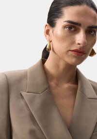 Mujer con el cabello oscuro peinado hacia atrás, vistiendo una chaqueta beige y grandes pendientes de aro dorados, contra un fondo claro y liso.