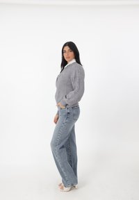 Grauer Kabelstrickpullover mit hohem Kragen, kombiniert mit hochtaillierten hellblauen Jeans. Das Modell steht seitlich und zeigt die lockere Passform und den strukturierten Stoff.