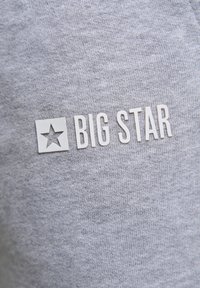 Pilkas audinys marškinėliai su balta logo, kurioje pavaizduota žvaigždė ir tekstas "BIG STAR" išryškintais, storais kiekvienos raidės rašmenimis, sukuriantys tekstūrinį kontrastą.