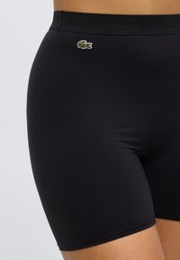 Shorts en tissu noir à texture lisse, avec une large ceinture noire et un petit logo crocodile vert brodé sur le côté.