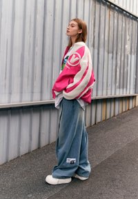 Chaqueta de forro polar rosa oversized con rayas blancas, combinada con pantalones vaqueros azules holgados que presentan una etiqueta. El modelo lleva zapatillas blancas. Vista de perfil.