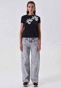 Zwarte T-shirt met witte bloemenborduursel, gecombineerd met lichtgrijze wijde jeans. Sneakers hebben zwarte, witte en tan accenten.