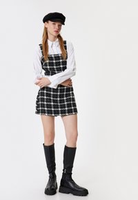 Vestido de cuadros en blanco y negro con una tela texturizada, superpuesto a una camisa blanca con cuello, combinado con botas negras hasta la rodilla y una gorra a juego.