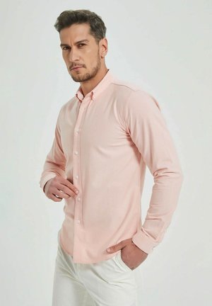 Chemise longue à manches roses clair avec fermeture à boutons et col pointu. Le tissu semble lisse ; conception ajustée avec des manches à poignets.