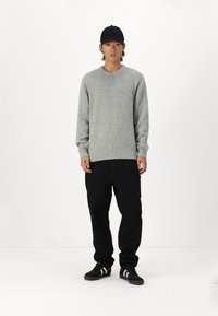 Pelēks adīts džemperis ar ribotām piedurknēm un apakšmalu, kopā ar melniem brīviem biksēm un melnām sporta apaviem ar baltām svītrām.