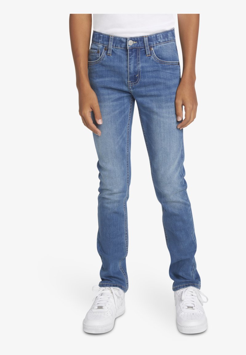 Jeans de mezclilla azul ajustados con un lavado desgastado, diseño de cinco bolsillos y costuras en contraste. Llevados con zapatillas blancas.