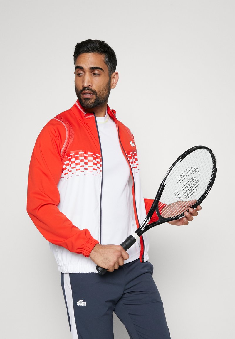 Giacca da pista rossa e bianca con un motivo a scacchi, abbinata a una maglietta bianca e pantaloni blu navy, mentre tiene una racchetta da tennis nera.