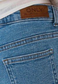 Blauwe spijkerbroek van denim met een bruin leren label met de tekst "ONLY Blue Denim." Voorzien van gestikte zakken en een vervaagde afwerking.