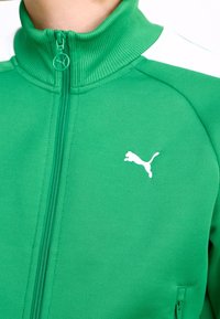 Veste zippée verte en tissu texturé avec un col montant, ornée d'accents blancs et d'un petit logo Puma blanc sur le devant.