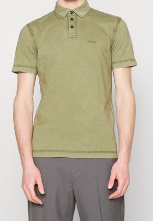 Mand iført olivengrøn kortærmet polo med tre sorte knapper og subtilt logo på brystet, kombineret med grå bukser.