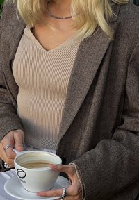 Femme blonde portant une veste marron en chevrons et un haut côtelé beige tient une tasse à café blanche avec une soucoupe.