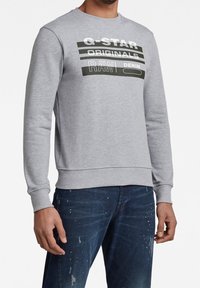 Felpa grigia con collo a giro, con grafiche testuali in bianco e nero a contrasto. Il testo include "G-STAR ORIGINALS RAW DENIM."