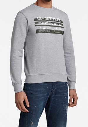 Grå sweatshirt med rund hals, med kontrasterande svart och vit textgrafik. Texten inkluderar "G-STAR ORIGINALS RAW DENIM."