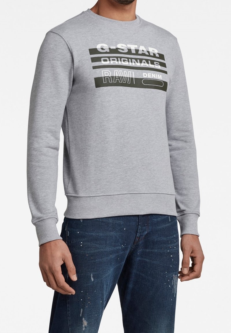 Felpa grigia con collo a giro, con grafiche testuali in bianco e nero a contrasto. Il testo include "G-STAR ORIGINALS RAW DENIM."