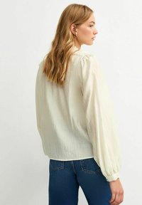 Blusa de color crema con tejido texturizado, mangas abullonadas y cuello con volantes, combinada con jeans ajustados azules.
