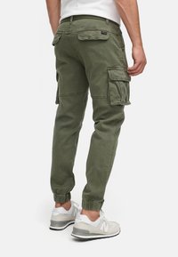Homme portant un pantalon cargo vert olive avec des poches à rabat et des baskets blanches New Balance, debout devant un fond blanc.