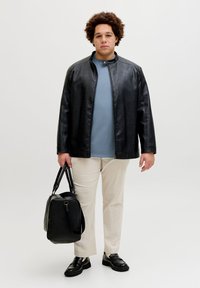 Άτομο με σγουρά μαλλιά που φοράει μαύρο δερμάτινο μπουφάν, μπλε πουκάμισο, μπεζ παντελόνι, μαύρα παπούτσια, κρατώντας μαύρη δερμάτινη τσάντα σε λευκό φόντο.