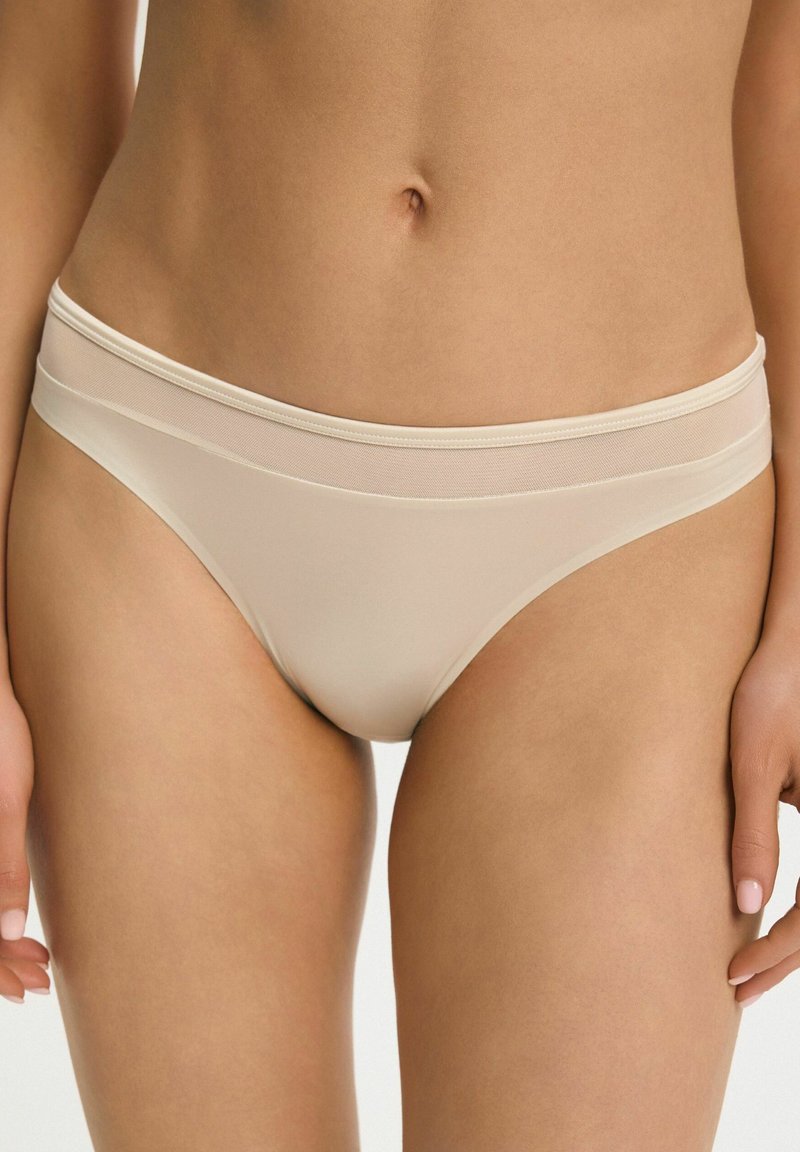 Kobieta w beżowej bieliźnie typu bikini z niskim stanem i przezroczystym, siateczkowym paskiem, widoczna od talii do górnej części ud.