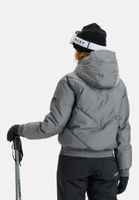 Giacca a vento grigia con cappuccio, caratterizzata da un motivo a scacchi e polsini elasticizzati, abbinata a pantaloni da neve neri e guanti. È indossato un berretto nero.