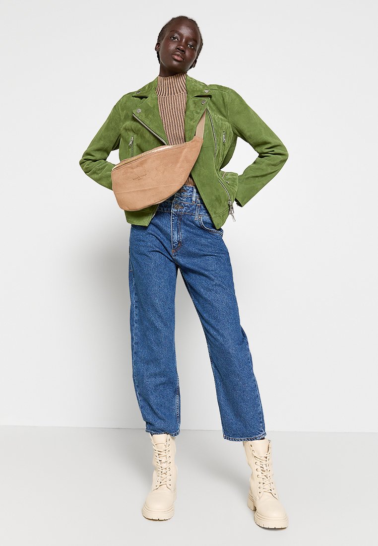 Femme portant une veste en daim verte, un col roulé rayé brun, un jean bleu, des bottes beiges à lacets et un sac bandoulière beige, se tenant devant un fond blanc.