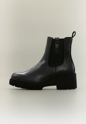 Bottine en cuir noir avec semelle épaisse, panneau latéral élastique, languette de tirage et bouton métallique décoratif sur fond neutre.
