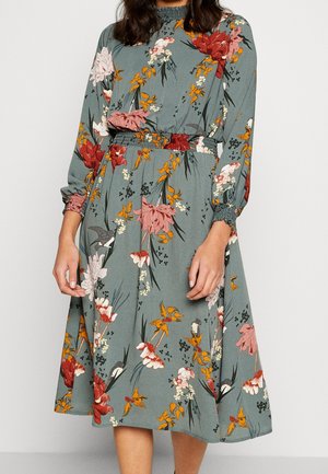 Robe fleurie en bleu-vert atténué avec des manches longues, taille smockée et motifs de fleurs colorés en rouge, rose et orange sur un fond vert.