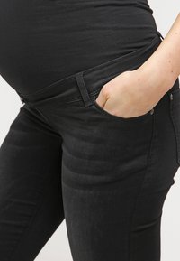 Jeans de maternidade pretos com um design ajustado e cintura elástica, apresentando bolsos frontais e uma textura suave.