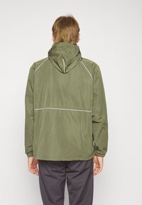 Olive green windbreaker with a hood, featuring reflective white accents, a relaxed fit, and elastic cuffs. Back view shows smooth texture.