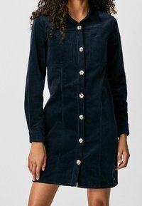 Marinblå manchesterkjol med långa ärmar, knappstängning fram, två bröstfickor och silverfärgade knappar. Slät textur och skräddarsydd passform.