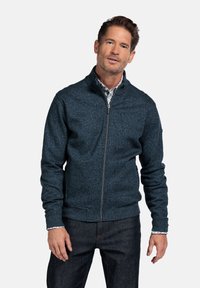 Marineblaue strukturierte Zip-Jacke mit Stehkragen, die mit gerippten Bündchen und Saum versehen ist. Sie wird über einem gemusterten Hemd und dunklen Jeans getragen.