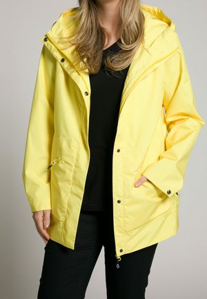 Personne portant un imperméable jaune vif par-dessus une chemise noire et un pantalon noir, une main dans la poche du manteau et les cheveux lâchés visibles.