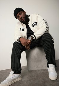 Vit basebolljacka med svarta bokstäver, svarta randiga byxor och vita sneakers. Har en svart stickad mössa och en silverfärgad klocka.