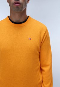 Maglione arancione lavorato a maglia con scollo rotondo e un piccolo emblema della bandiera norvegese sul petto. Texture liscia, design aderente.