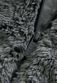Chlpatá sivá fleecová látka s výraznou textúrou, s čiernym zipsom s označením "BAKER" v detailnom zábere.