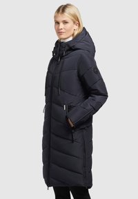 Mantou lung puffer de culoare bleumarin, cu un design capitonat, glugă ajustabilă, fermoar frontal și buzunare laterale. Material texturat cu un finisaj mate.