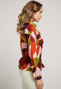 Chemise à motifs colorés présentant des formes géométriques en rouge, rose, vert et marron. Modèle à boutons avec un col pointu et des poignets.