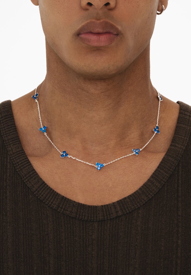 Silberne Kettenhalskette mit blauen Edelsteinen und blütenartigen Designs, getragen am Hals einer Person über einem dunkelbraunen Kleidungsstück.