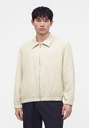 Homme portant une veste beige clair zippée avec col et poignets élastiques, assortie à un pantalon foncé, debout devant un fond blanc uni.