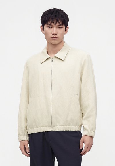 Homme portant une veste beige clair zippée avec col et poignets élastiques, assortie à un pantalon foncé, debout devant un fond blanc uni.