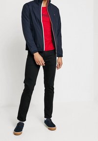 Mand iført en marineblå bomberjakke over en rød grafisk T-shirt, sorte slim-fit bukser og marineblå sneakers med hvide såler, der står mod en hvid baggrund.