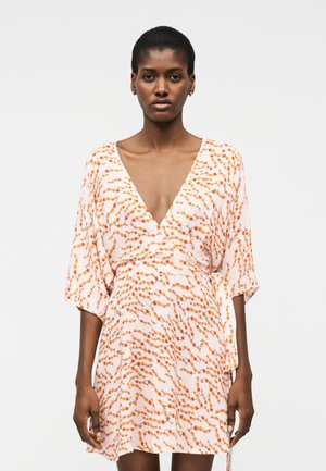 Γυναίκα που φοράει λευκό φόρεμα τύπου wrap με πορτοκαλί λουλουδάτο μοτίβο και κοντά, φαρδιά μανίκια, στέκεται μπροστά σε απλό ανοιχτό φόντο.