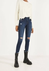 Persona vestida con un suéter corto blanco, jeans ajustados oscuros con rotos, y botas de plataforma negras gruesas, de pie sobre un suelo blanco.