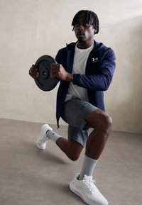 Sort vægplade holdt af en person i en navy jakke og hvid t-shirt, der udfører en lunge iført grå shorts og hvide sneakers. Tekstureret gulv.