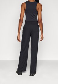 Kobieta stoi tyłem, ubrana w czarną bezrękawnik, wysokogatunkowe czarne spodnie z szerokimi nogawkami i czarne sneakersy.