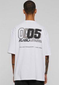 Balts kokvilnas T-krekls ar lielu grafiku, kurā redzami skaitļi "005" un logo "FUBU" melnā un pelēkā krāsā uz muguras. Īsas piedurknes.
