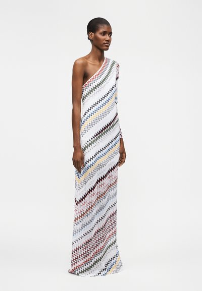 Γυναίκα που φοράει φόρεμα ενός ώμου, μάκρος μέχρι το πάτωμα, με διαγώνιες ζιγκ-ζαγκ ρίγες σε πολλαπλά χρώματα σε λευκό φόντο.