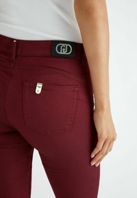 Femme portant un pantalon ajusté bordeaux avec une poche arrière et un patch logo, associé à un haut blanc côtelé, main reposant le long du corps.